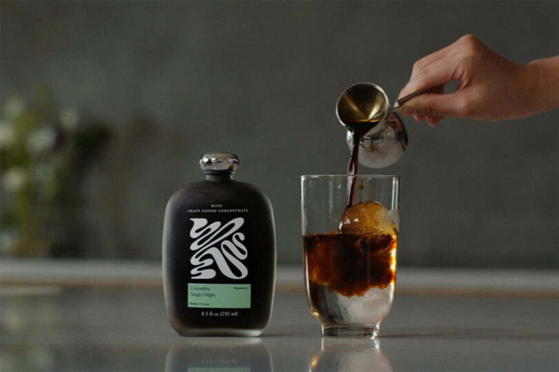 Dark craft coffee concentrate being poured from a metal cup into a glass filled with ice.