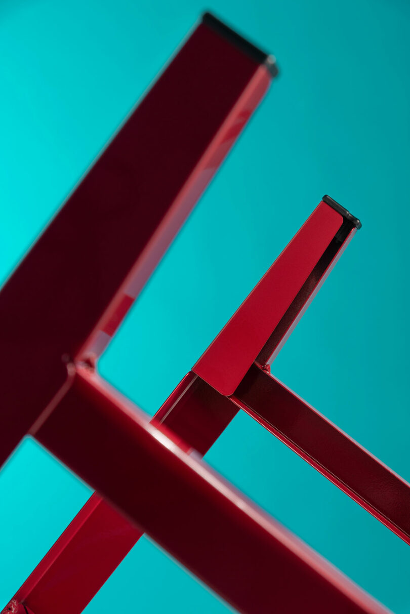 Close-up of the red metal legs of the Bishop Chair against a turquoise background, showing the precise welding.