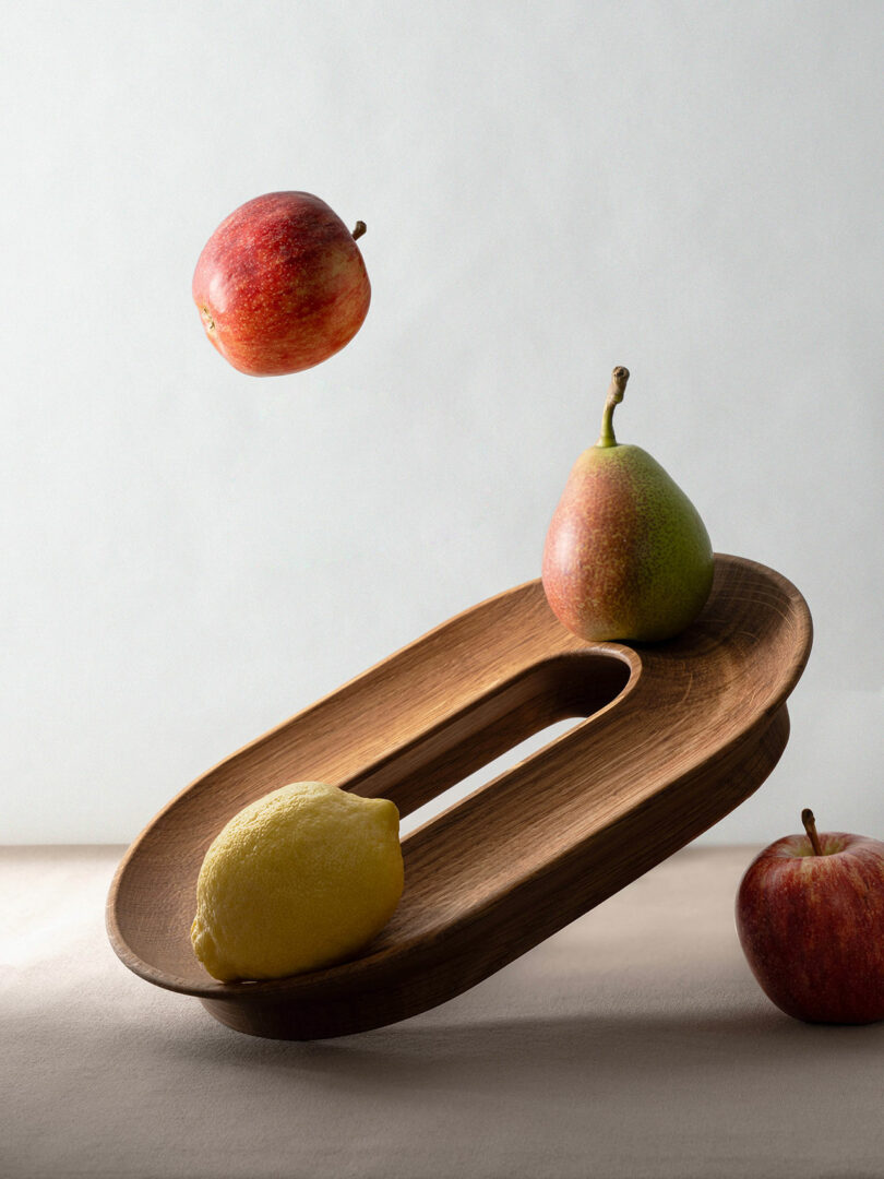 An apple floating above a wooden tray with a lemon and pear nearby on a neutral background.