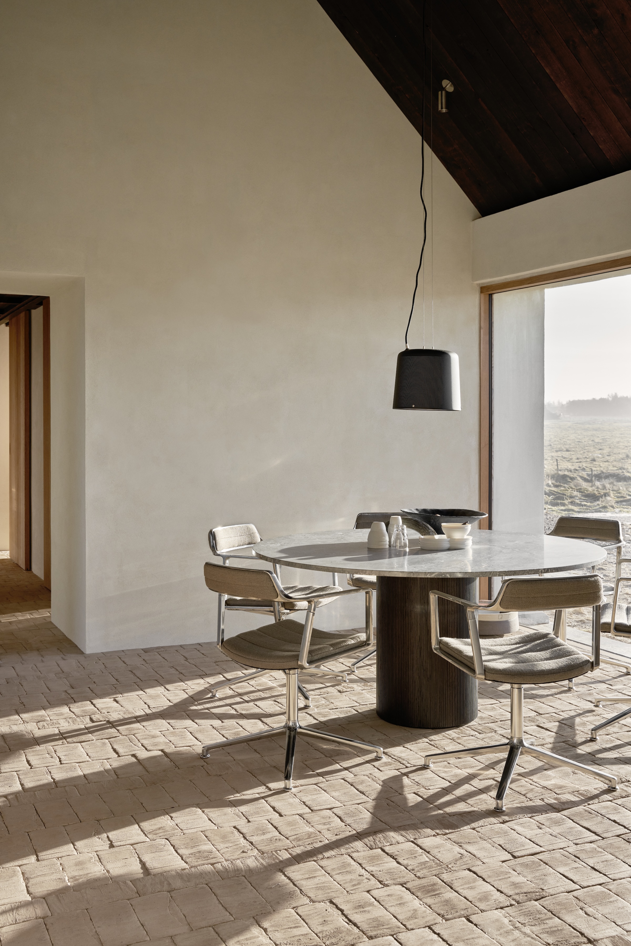 A round limestone-topped dining table with black swivel chairs and a perforated pendant light.