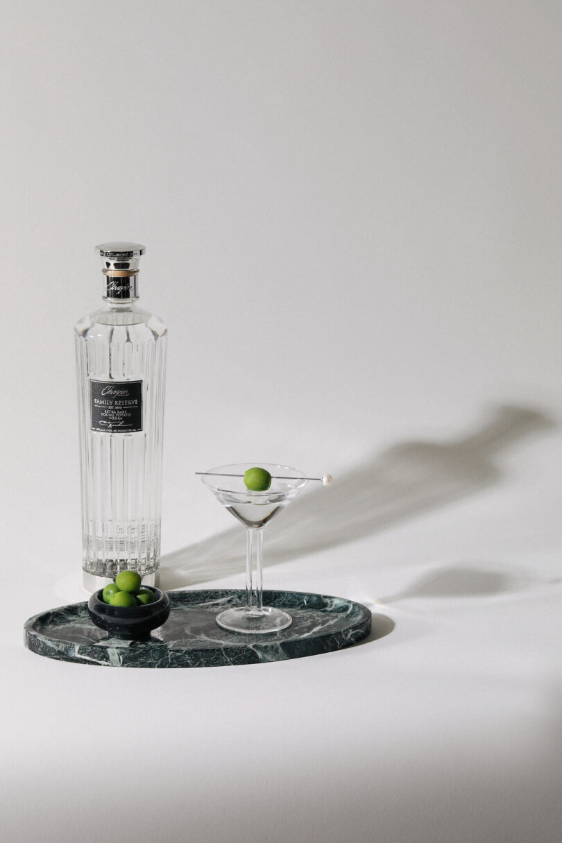 A bottle and martini glass set styled on a dark green marble tray with a side of olives.