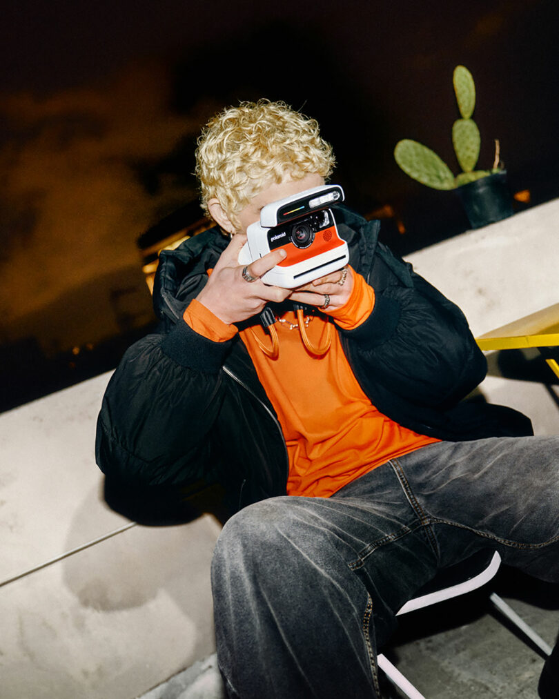 A person using a Polaroid camera at night outdoors, with a cactus and soft lighting in the background.