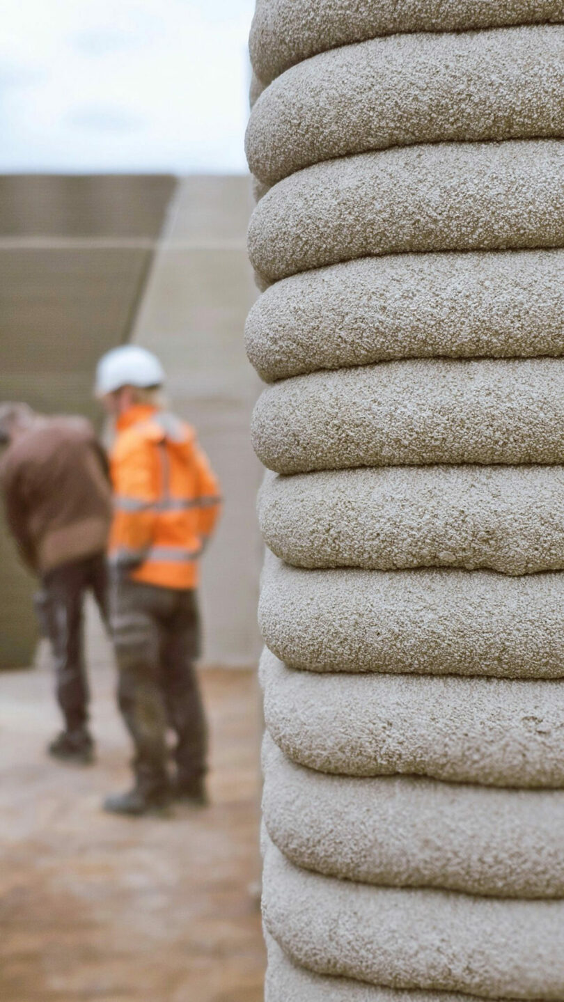 Close-up of the horizontal layers of 3D-printed concrete with construction workers in the background.