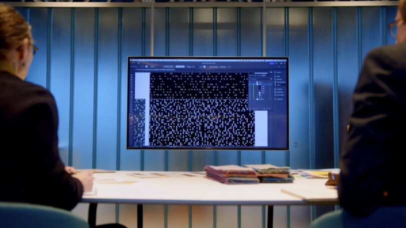 A designer works at a computer screen displaying digital patterns with fabric swatches on the desk.