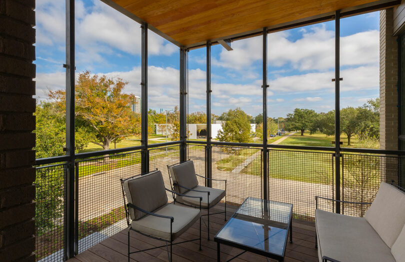 A screened balcony with comfortable seating looking out over green trees and a modern building.