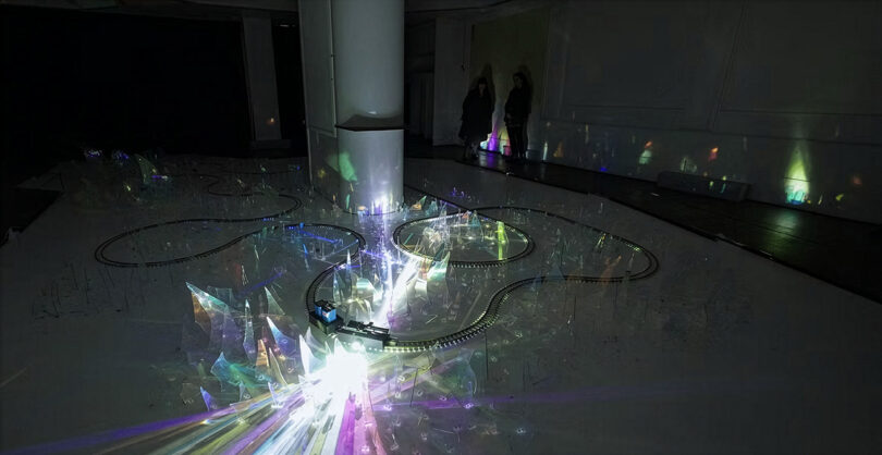 An installation featuring light reflecting off crystal-like structures in a dark room with human silhouettes in the background.