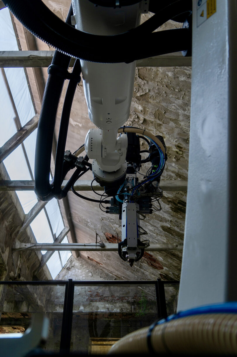 A large industrial robotic arm suspended from a ceiling in an old building with a glass roof.