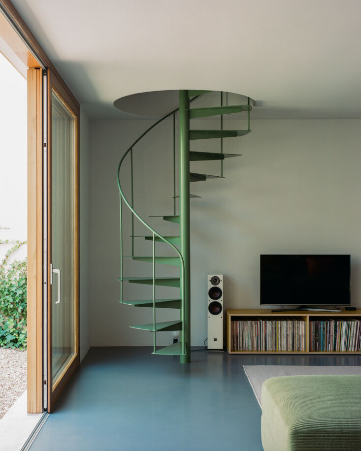 A sculptural green steel spiral staircase winding up from a light blue industrial floor.