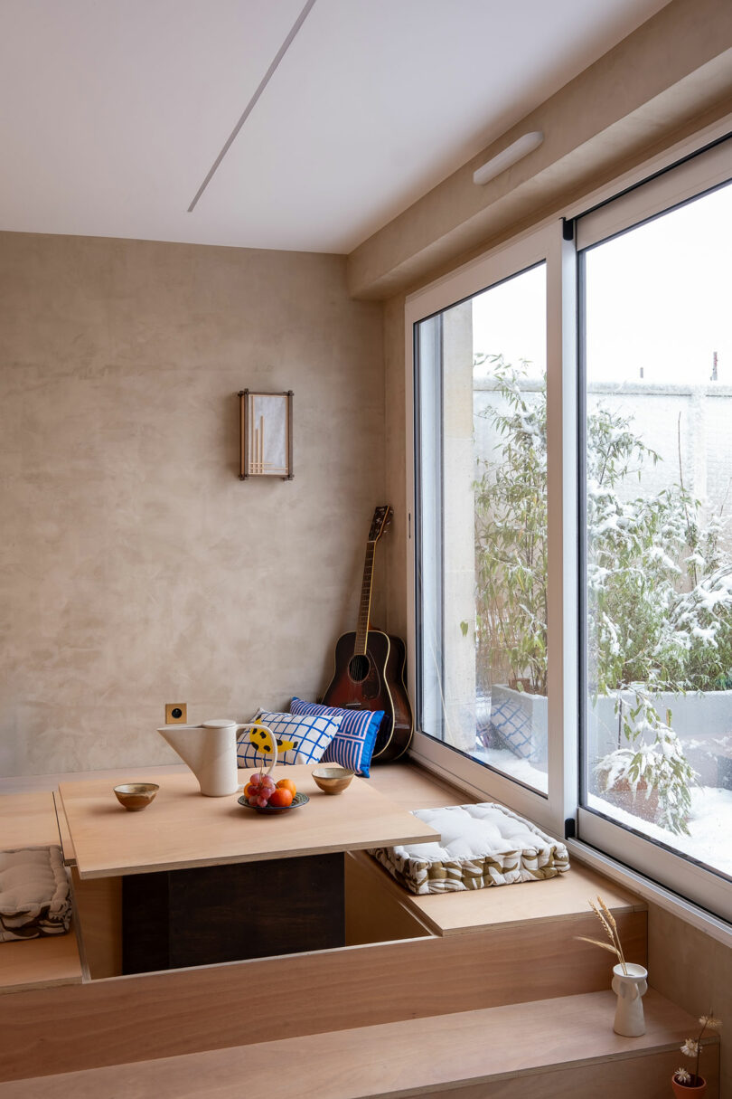 A sunken wooden table within a platform featuring cushions and a guitar by a snowy window.