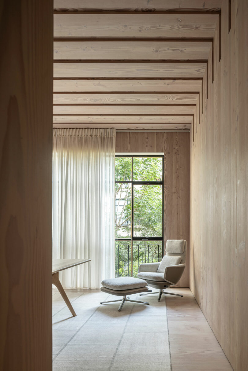 An armchair and ottoman placed next to a window with sheer curtains, showing a connection to outside greenery.