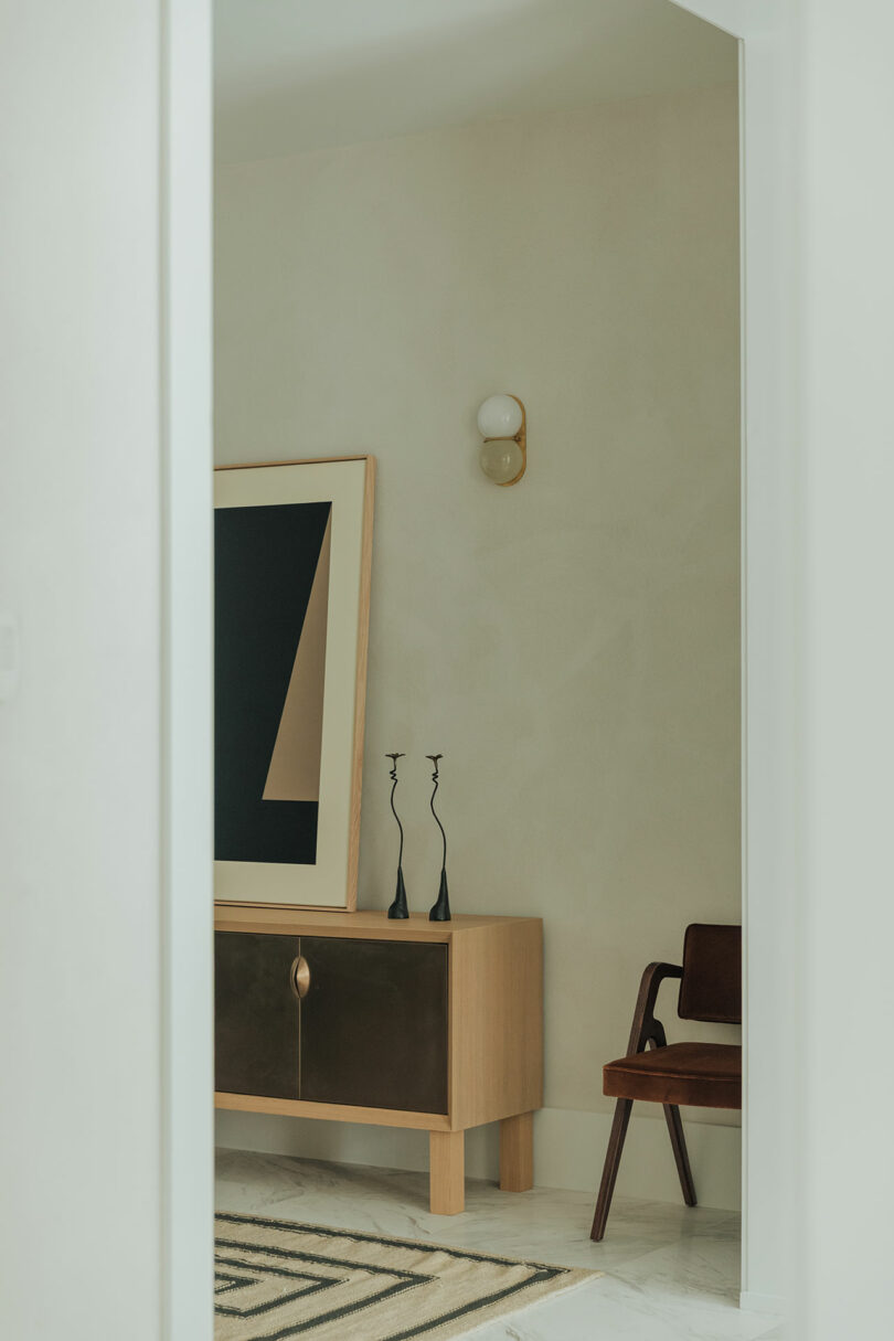 A minimalist wooden cabinet with abstract art, black candlesticks, and a single wooden chair.