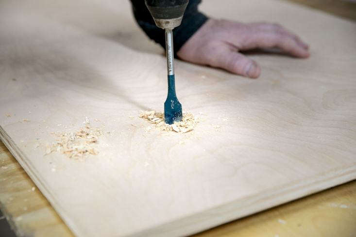 A person using a power drill held strictly perpendicular to a wooden board.