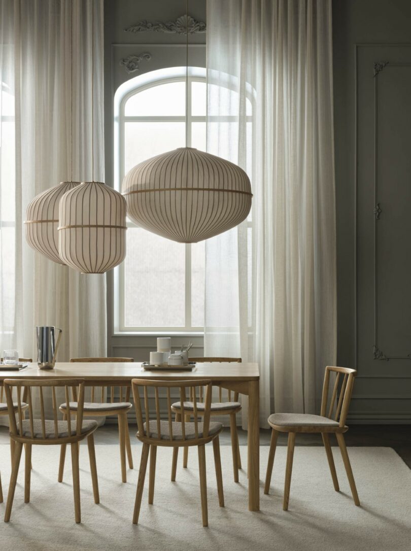 A minimalist dining room with four chairs and a wooden table set against large arched windows and sheer curtains.