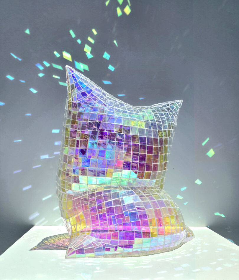 A sculpted pillow covered in holographic mosaic tiles reflecting rainbow light onto a white surface.