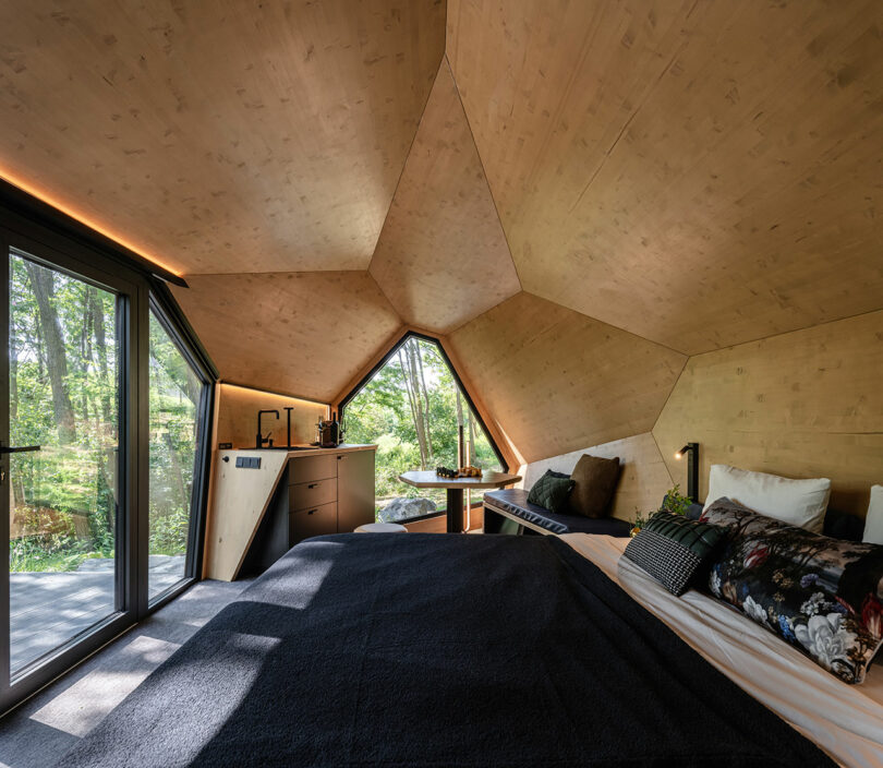 Modern cabin interior showing the bedroom area, small dining table, and large window overlooking the woods.