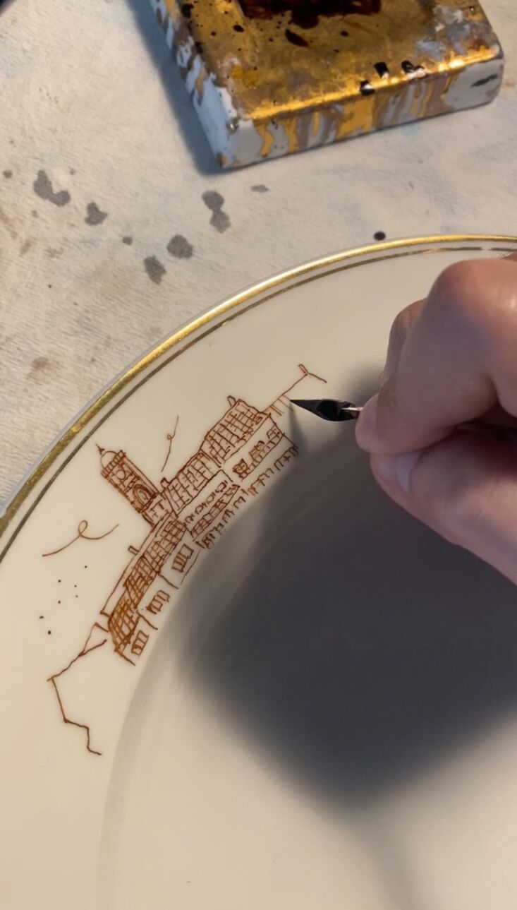 Close-up of a hand applying gold glaze to a porcelain plate using a dip pen.