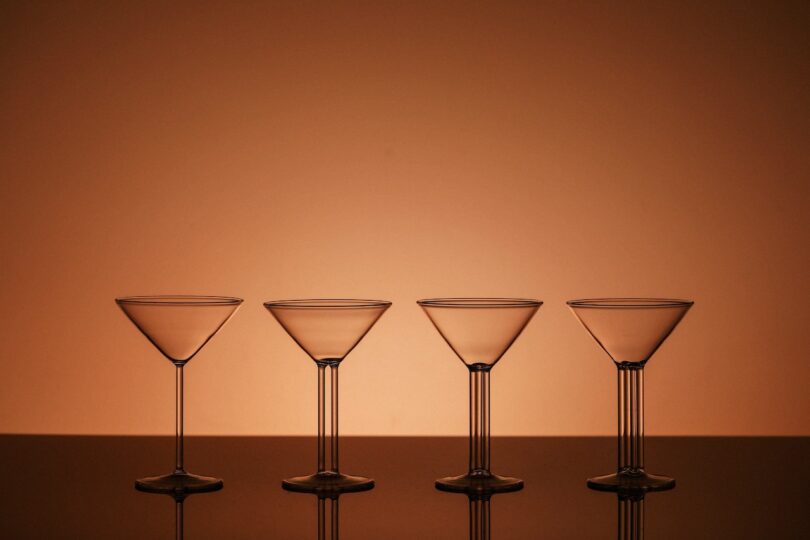 Four empty Ernestine mini martini glasses aligned against an orange gradient background, showing different stem designs.