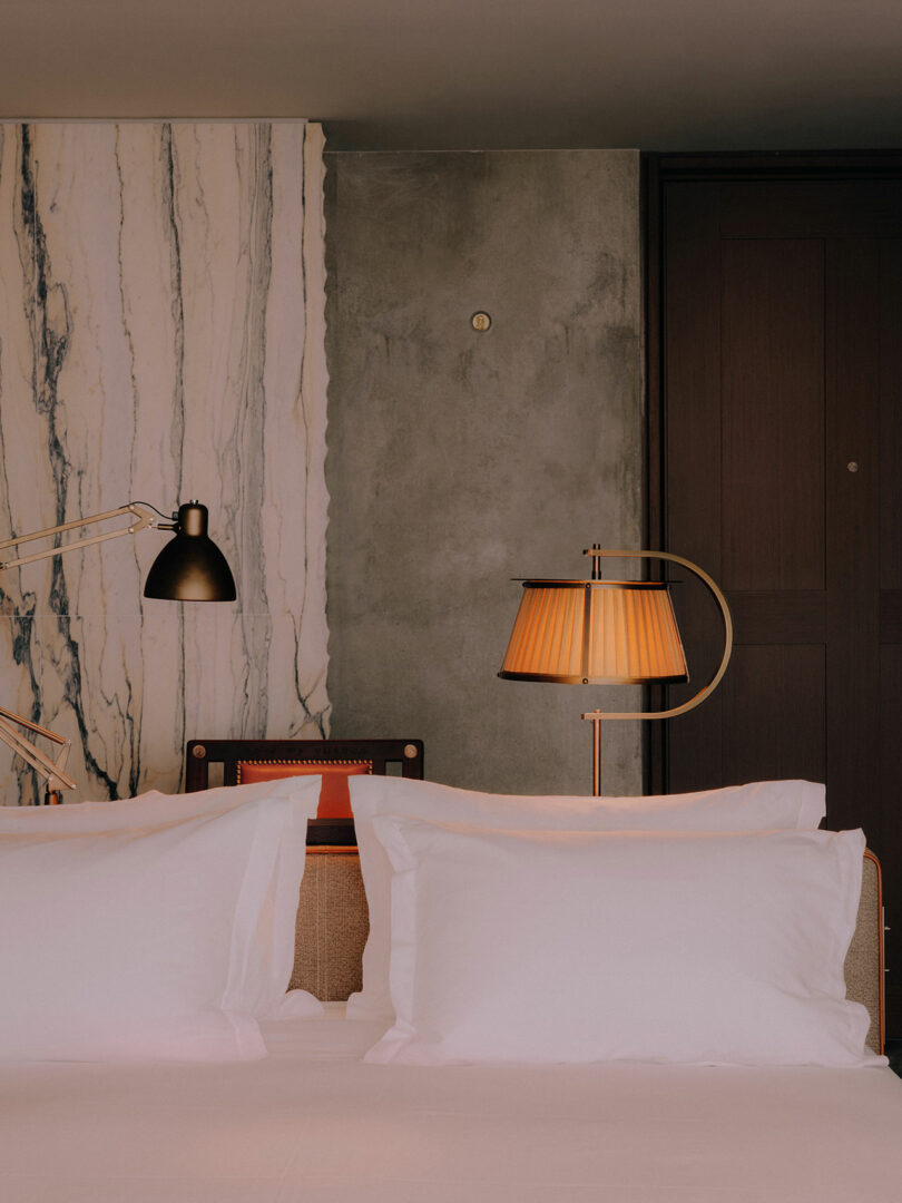 A neatly made bed with white pillows against a backdrop of marble and raw concrete walls.
