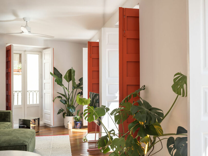A sunlit living room with white walls, wooden floors, large green plants, and bright red balcony doors.