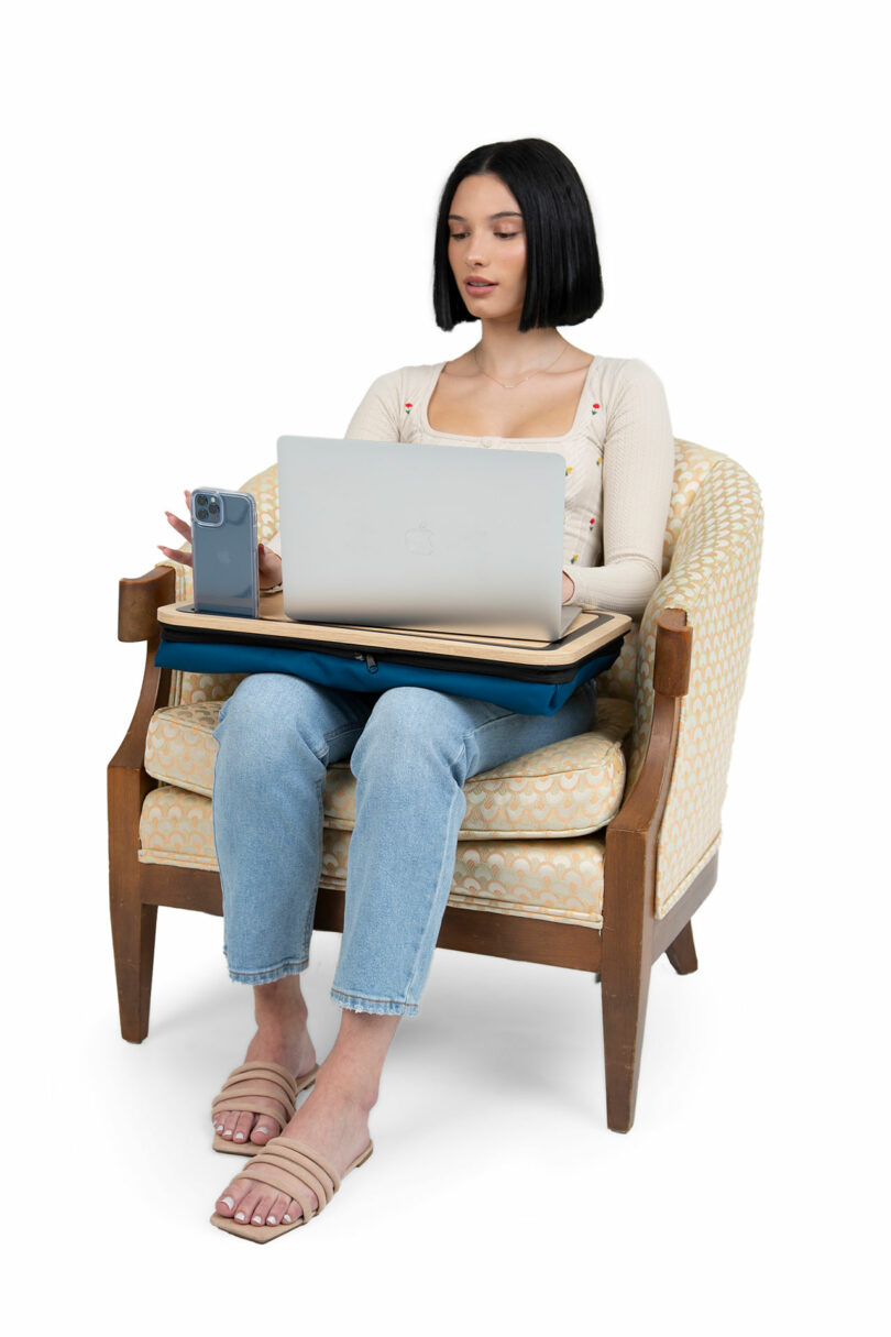 A woman using a laptop on the lap desk while sitting in a comfortable armchair, with her phone docked vertically.
