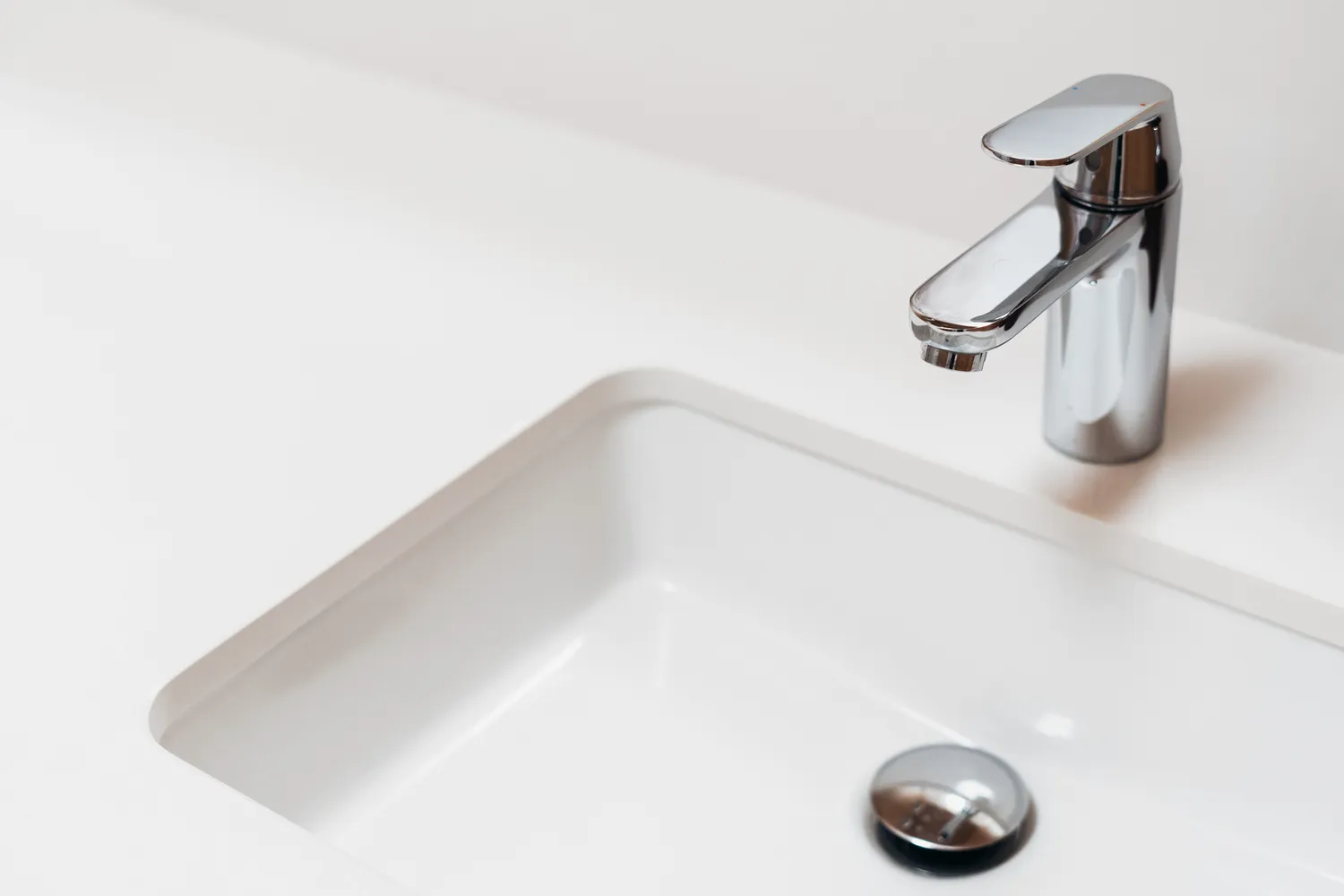Close-up of a minimalist white bathroom sink with a chrome single-handle faucet and pop-up drain mechanism.