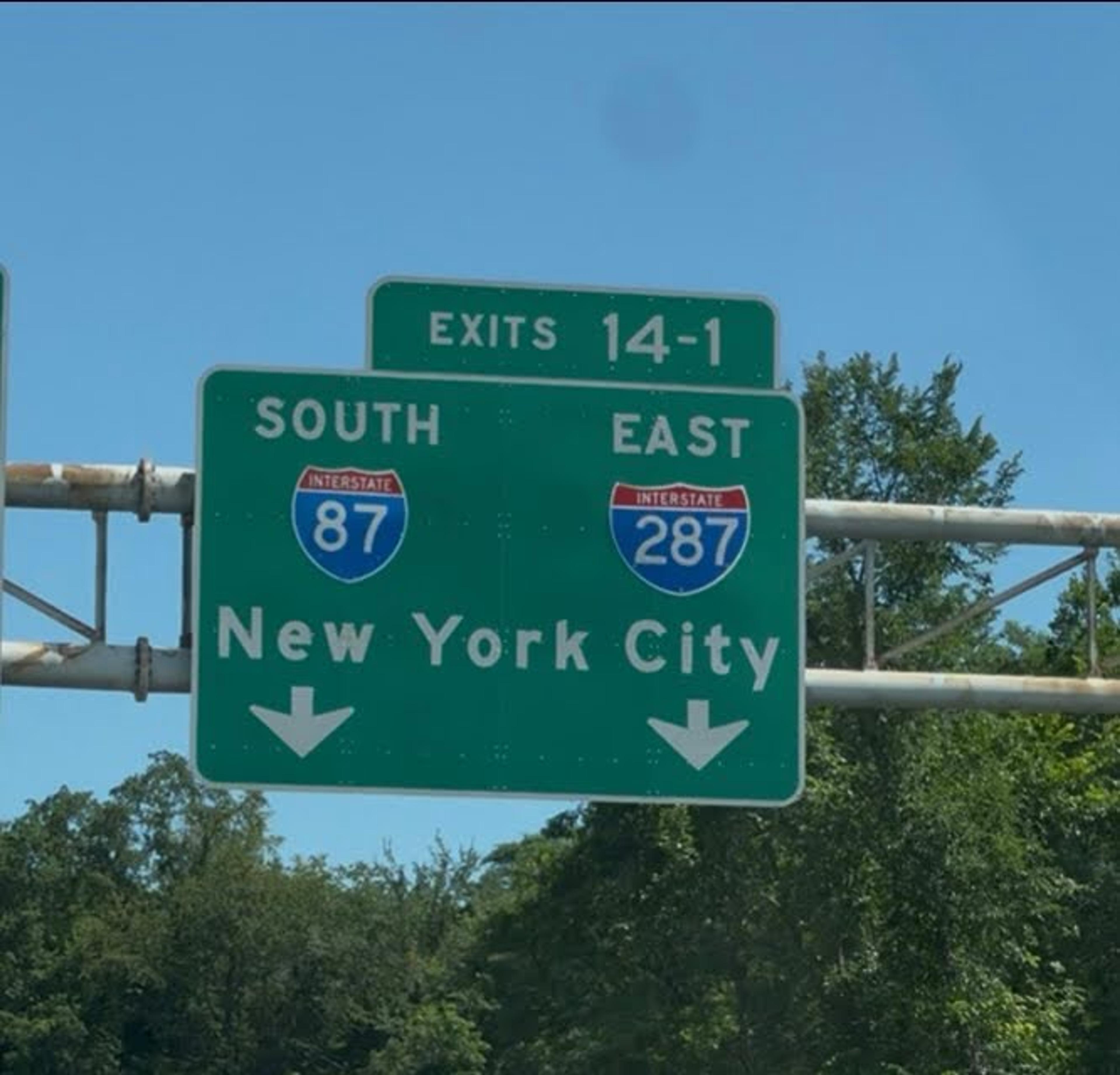 A green highway sign indicating the direction to New York with lush trees in the background.