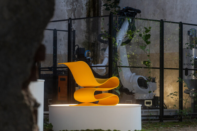 A bright yellow curved 3D-printed chair in front of a robotic arm.
