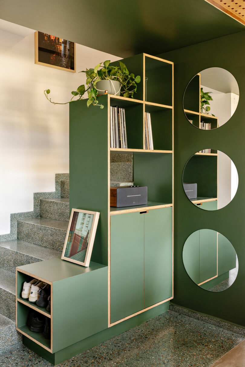 Modern green entryway storage unit with circular mirrors and shoe cubbies next to a staircase.