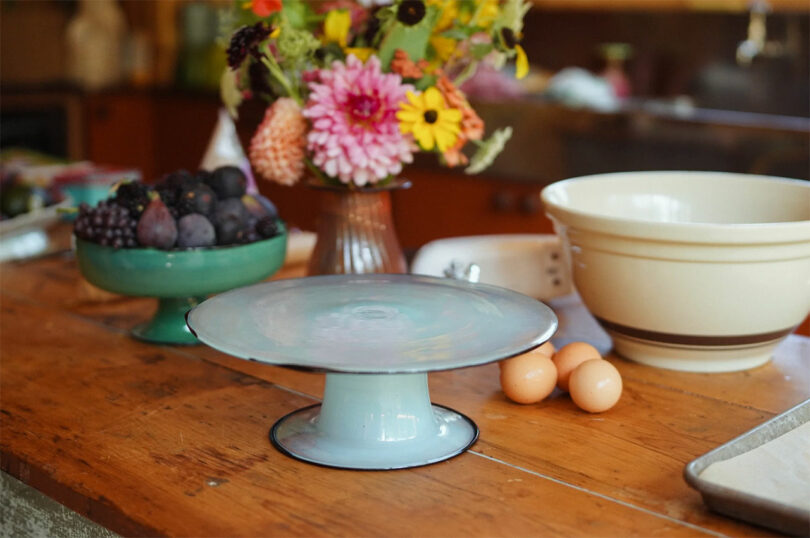 A glass cake stand and kitchen essentials arranged on a rustic wooden countertop.