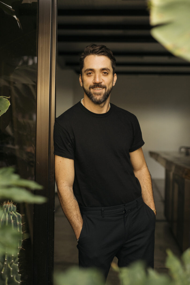 Portrait of designer Diego Olivero in a casual black outfit standing near green plants.