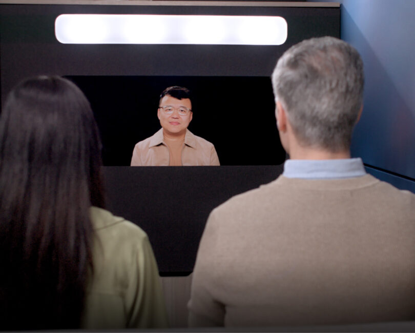 Two office workers engaged in a life-size video call via the Ocular View, showing natural eye contact with an Asian man on the screen.