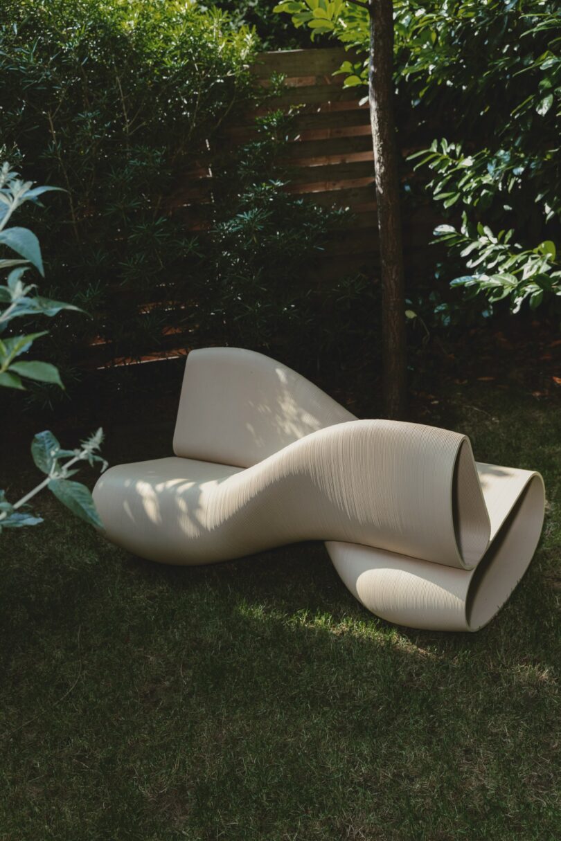 A sculptural beige lounge chair sitting on a lush green lawn surrounded by trees.