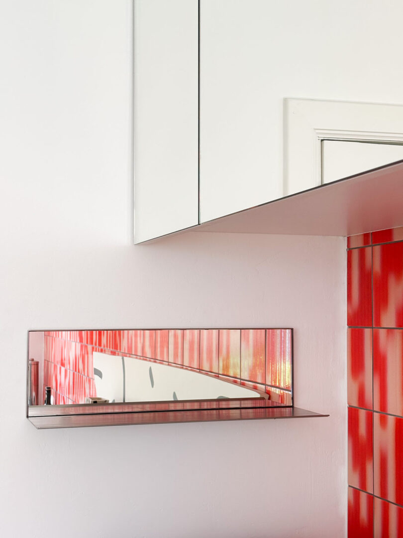 A wall-mounted mirror shelf reflecting a red and white patterned wall in a modern room.
