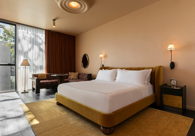 A hotel guest room featuring a mustard-yellow headboard, matching rug, and large windows with layered curtains.