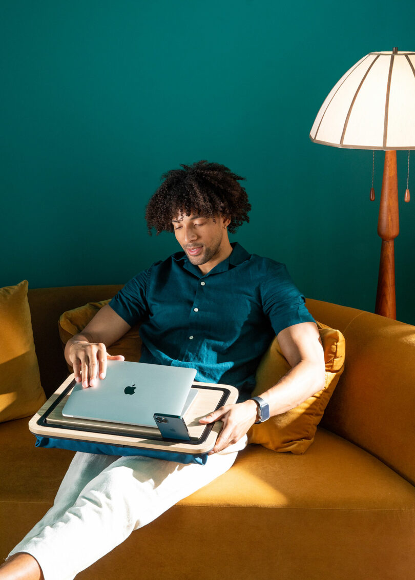 A man using multiple apple devices while sitting on a mustard yellow couch in a stylized living room.